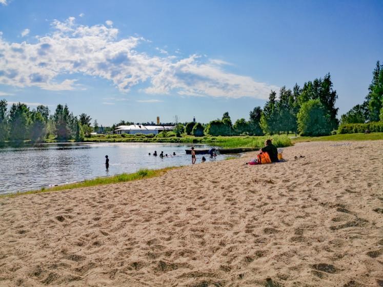 Lamminjärven uimaranta. Etualalla hiekkaranta, jossa ihmisiä ja laituri. Järvessä on ihmisiä uimassa. Taka-alalla puita. Sää on puolipilvinen. 