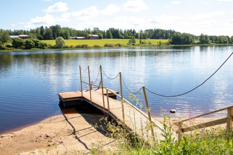 Hebrominrannan uimaranta. Etualalla laituri. Tyyni järvi. Taka-alalla peltoa ja metsää. 