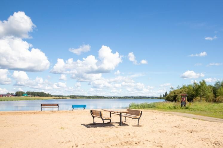 Hirvijärven uimaranta. Etualalla hiekkaranta, jossa pöytäryhmä ja kaksi penkkiä. Taka-alalla on tyyni järvi. Vastarannalla on maatila, peltoa ja puita. Sää on puolipilvinen.