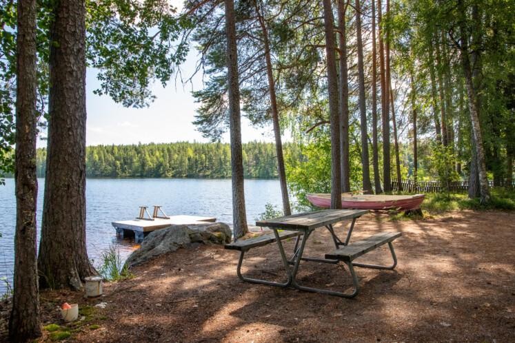 Kolhonjärven uimaranta. Etualalla pöytäryhmä metsäisellä maalla. Taka-alalla puita, laituri ja tyyni järvi. Sää on aurinkoinen. 