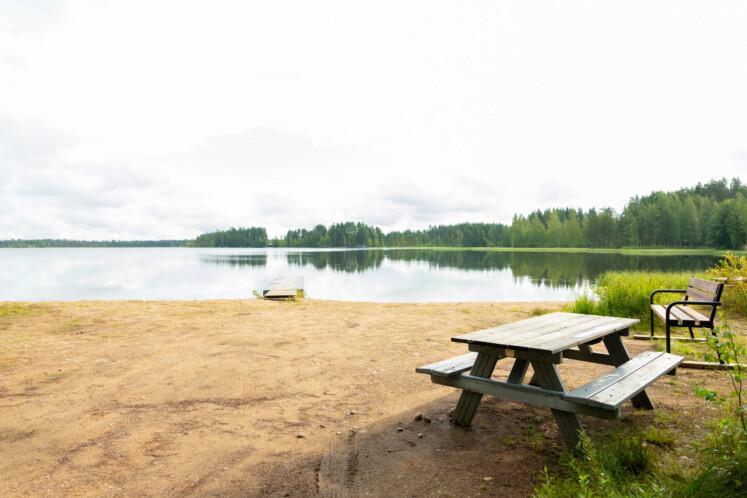 Korvajärven uimaranta. Etualalla hiekkaranta, jossa pöytäryhmä ja penkki. Taka-alalla laituri, tyyni järvi ja metsää. Sää on pilvinen. 