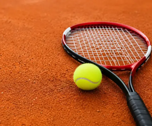 Tennismaila ja tennispallo oranssilla massakentällä. Lähikuvassa maila lepää kentän pinnalla pallon vieressä.